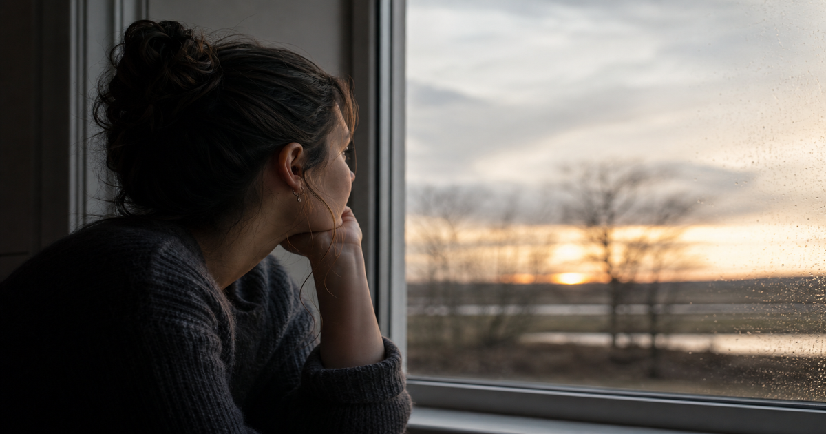 Iemand kijkt peinzend uit het raam bij zonsondergang — reflectie en terugkeer na transitie